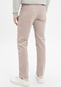 Man in beige corduroy broek en witte sneakers, staand tegen een effen witte achtergrond, gezien vanaf de achterkant.