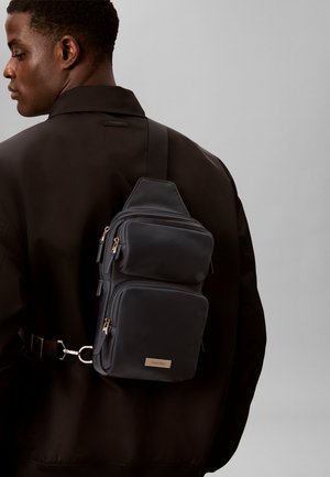 Mann trägt schwarze Jacke mit schwarzer Mehrfach-Taschen-Umhängetasche, die diagonal über seinen Rücken vor einem einfarbigen grauen Hintergrund getragen wird.