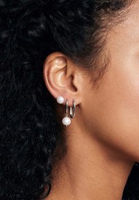 Srebrne kolczyki w kształcie obręczy z wiszącą białą perłą, charakteryzujące się polerowanym wykończeniem i gładką teksturą. Dwa różne rozmiary pereł tworzą kontrast.
