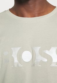 Detailní záběr na krk a horní část hrudníku v lehce béžové košili se stříbrným reflexním logem "BOSS" přes hrudník.