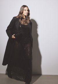 Largo vestido negro con superposiciones transparentes, adornado con sutiles lentejuelas. Combinado con una chaqueta negra y un clutch, con un diseño estructurado.