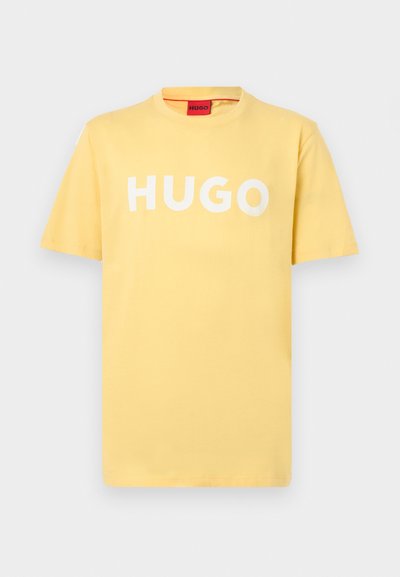 Жълта тениска с къси ръкави и кръгло деколте, с голям бял надпис "HUGO" отпечатан на гърдите и червен бранд етикет вътре в яката.