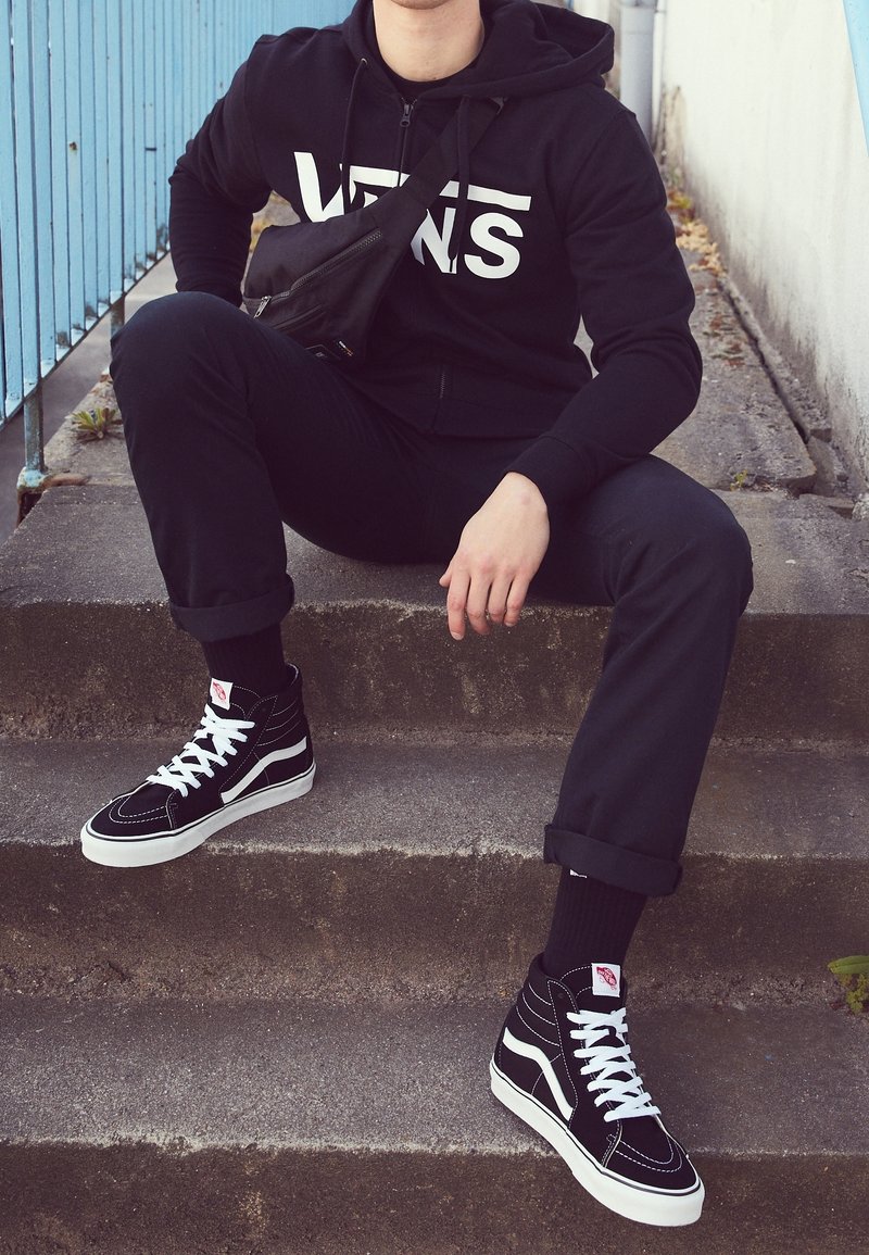 Personne portant un sweat à capuche Vans noir, un pantalon noir et des baskets montantes Vans noires, assise sur des marches en béton avec une rambarde bleue à proximité.