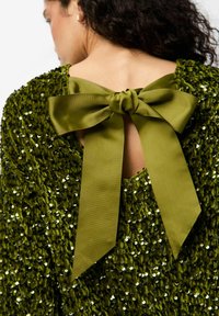 Smaragdzaļa, sekvīnām rotāta blūze ar izgriezumu aizmugurē. Satur lielu satīna loku no atbilstošas zaļas auduma, pievienojot tekstūru un detaļas.