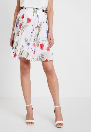 Frau trägt einen weißen Blumensrock mit roten, lila und grünen Blumen, kombiniert mit weißen High-Heel-Sandalen.