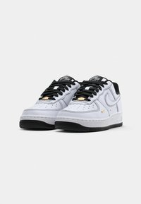 Hvite Nike Air Force 1-sneakers med svarte såler, svarte snører og små gullswoosh-detaljer på siden, vist fra frontvinkel.