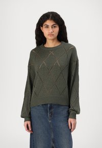 Grüner gestrickter Pullover mit Rautenausschnitten, einem gerippten Halsausschnitt und langen Ärmeln, kombiniert mit einem Jeansrock. Texturiert und lässiger Schnitt.