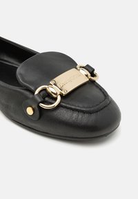 Mocassin en cuir noir avec plaque en métal doré et détails en anneaux sur la partie supérieure avant de la chaussure.