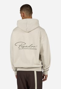 Kremfarget hettegenser med snøre i hetten, med en stor grå tekstlogo på baksiden. Mykt stoff med avslappet passform og klassisk design.