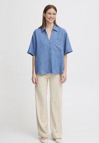 Camicia blu a maniche corte con bottoni e taschino, realizzata in tessuto leggero. Abbinata a pantaloni a gamba ampia color crema e scarpe beige.