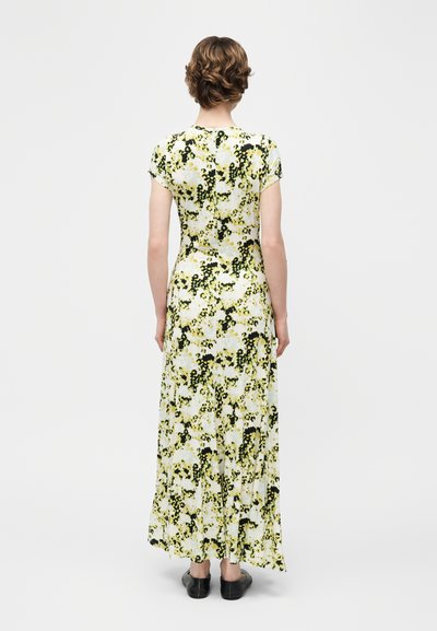 Γυναίκα με κοντά μαλλιά, ντυμένη με μακρύ φόρεμα με κοντά μανίκια και λουλουδάτο μοτίβο, φορόντας μαύρα παπούτσια, στέκεται με την πλάτη προς το μέρος μας σε λευκό φόντο.