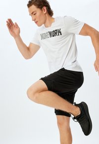 Hvid kortærmet skjorte med "MORE WORK" trykt i sort, parret med sorte atletiske shorts og sorte løbesko, i en dynamisk positur.