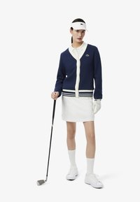 Cardigan blu navy con dettagli crema, polo bianca e gonna bianca. Completato con un visore, guanto da golf e sneaker bianche. Bastone in mano.