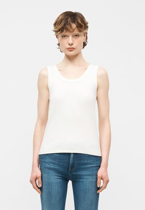 Jonge persoon met kort bruin haar, gekleed in een mouwloos wit topje en een blauwe spijkerbroek, staand tegen een effen lichte achtergrond.