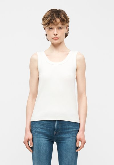 Jeune personne aux cheveux bruns courts portant un haut blanc sans manches et un jean bleu, debout devant un fond clair uni.