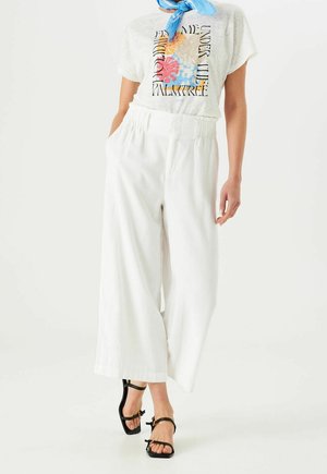 Person trägt weiße, weit geschnittene Hose, weißes T-Shirt mit buntem Design, blauen Halstuch und schwarze Riemchensandalen.