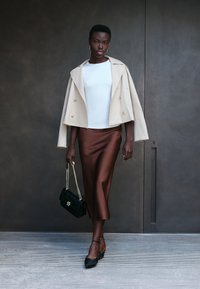 Giacca beige doppiopetto, top bianco, gonna midi in satin marrone. Borsa nera con tracolla a catena, scarpe nere con lacci alla caviglia, design elegante.