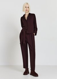Une chemise boutonnée bordeaux à manches longues associée à un pantalon à taille haute plissé et des bottes à mi-mollet, fabriqués dans un tissu lisse.