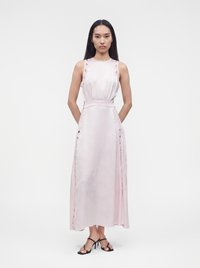 Femme debout portant une robe rose clair sans manches avec des détails en dentelle sur les côtés et des sandales à talons noires sur fond blanc.