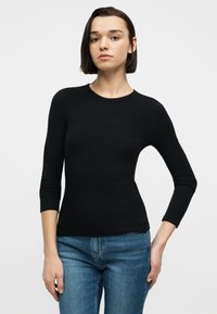 Νεαρή γυναίκα με κοντά σκούρα μαλλιά, φοράει μαύρο μακρυμάνικο εφαρμοστό μπλουζάκι και μπλε τζιν, στέκεται μπροστά σε ένα απλό λευκό φόντο.