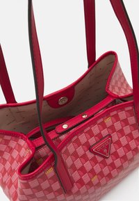 Sac à bandoulière rouge avec un motif à carreaux, logo à l'avant et deux longues anses. Dispose d'un intérieur doublé en tissu avec fermeture à pression.