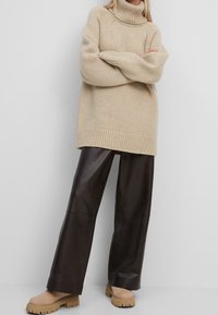 Beige neulottu pooloneule paksulla tekstuurilla, yhdistetty tummanruskeisiin leveälahkeisiin housuihin ja vaaleanruskeisiin korollisiin kenkiin.