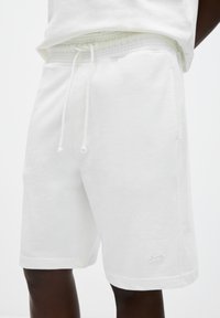 Shorts en coton blancs avec une taille élastique et un cordon de serrage. Comprend des poches latérales et un petit logo sur la jambe inférieure gauche. Texture douce et lisse.