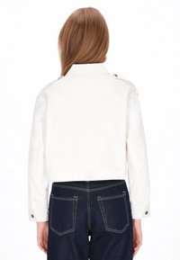 Witte cropped jas met een puntige kraag en knoopaccenten. Gecombineerd met donkere spijkerbroek met lichte stiksels op de zakken.