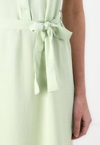 Rochie verde deschis dintr-un material texturat, cu o curea la talie legată și închidere cu nasturi, evidențiind margini netede și un design fluid.
