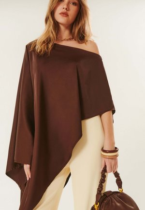 Femme portant un haut asymétrique marron épaule dénudée, pantalon crème, tenant un sac à main marron froncé avec des bracelets assortis au poignet.