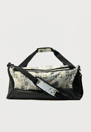 Zwarte duffelbag met een geometrisch patroon in groen, geel en wit. Heeft een zwarte riem en een handvat aan de bovenkant om te dragen.