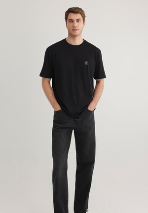Black T-shirt made of cotton with a round neckline, short sleeves, and a green logo on the chest. Worn with dark jeans.