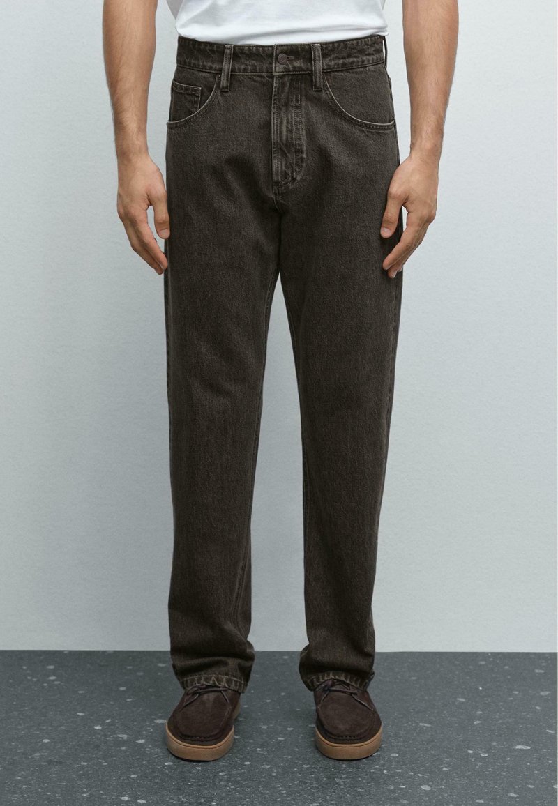 Mörkbruna denimbyxor med en avslappnad passform, femfickorsdesign och en synlig bältesöglor i midjan, ihop med bruna skor.