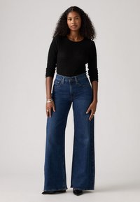 Jean taille haute à jambes larges en denim bleu foncé, associés à un haut noir texturé à manches longues. Les jeans sont dotés de poches latérales et d'une fermeture à bouton.
