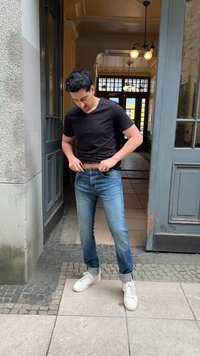 Άντρας με μαύρη μπλούζα και μπλε τζιν ρυθμίζει τη ζώνη του ενώ στέκεται έξω από την είσοδο ενός παλιού κτηρίου με πλακάκια στο δάπεδο και κρεμαστά φώτα.