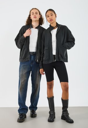Unisex mørkegrå hettejakke med glidelås, myk stoff, to frontlommer. Modellen har på seg svarte sykkelshorts og svarte knelange støvler med nagler.