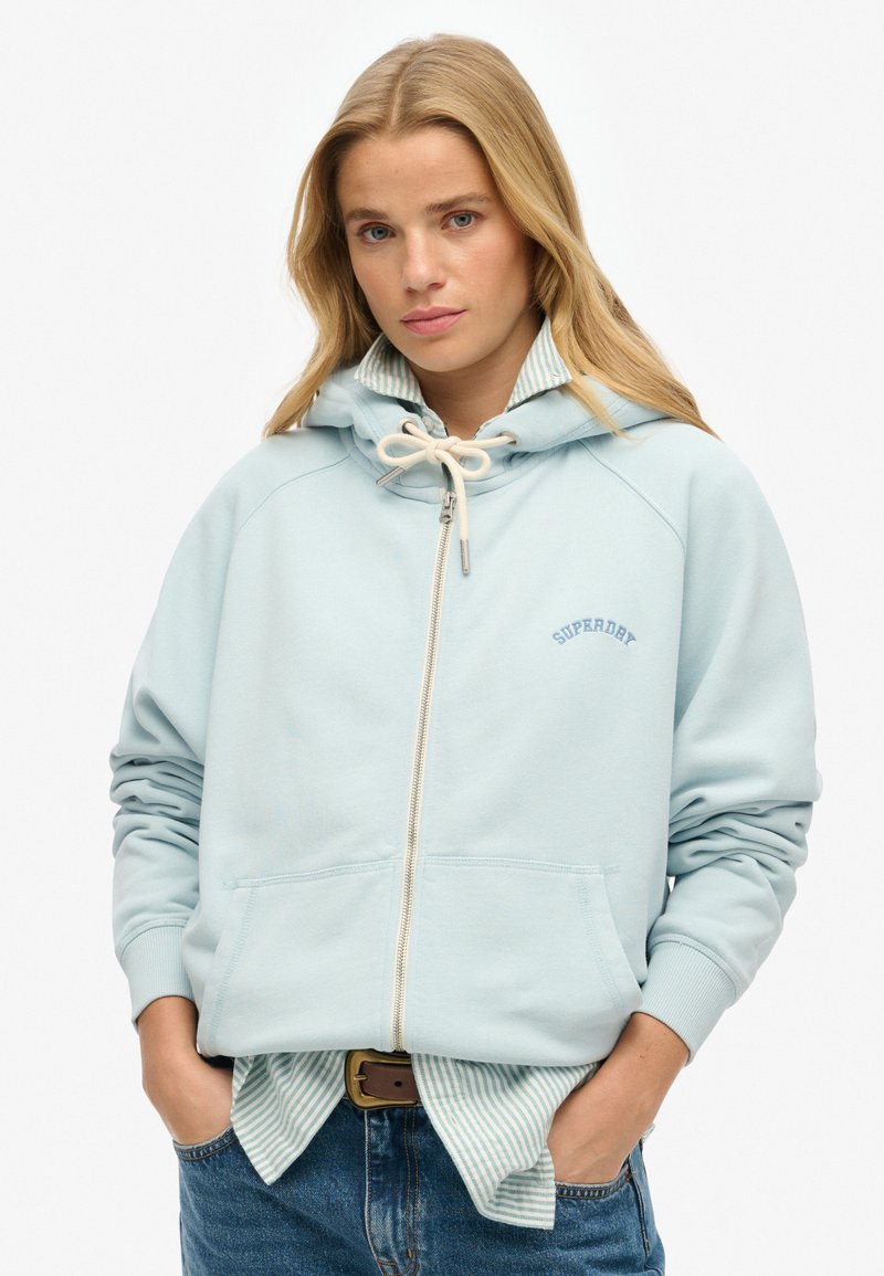 Felpa con zip di colore blu chiaro, dotata di cappuccio regolabile con cordino, tasca a marsupio e polsini a costine, indossata sopra una camicia a righe. Presenta il logo "SUPERDAY".