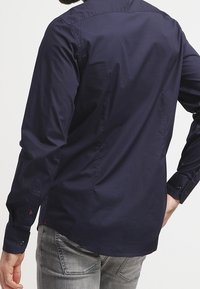 Navyblauw knoopshirt met lange mouwen, nauwsluitende pasvorm en een gladde textuur. Voorzien van manchetten met één knoop en een achterpanddetail.