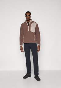 Pull en polaire marron avec un panneau de poitrine beige clair, fermeture zippée quart, deux poches latérales ; associé à un pantalon gris foncé et des chaussures de randonnée noires.