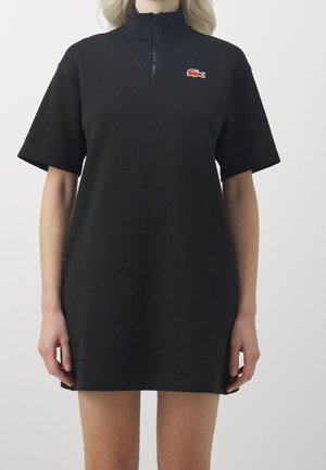 Frau trägt ein schwarzes Kurzarmkleid mit einem Viertel-Reißverschluss am Kragen und einem kleinen roten Krokodil-Logo auf der Brust.