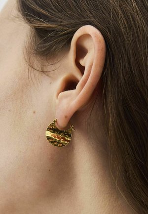 Primer plano de la oreja de una persona con cabello castaño, que lleva un pequeño pendiente de aro dorado martillado y redondo.