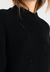 Maglione nero lavorato a maglia con collo alto, texture a costine e ritagli diagonali ai lati. Filato morbido e grosso senza decorazioni.