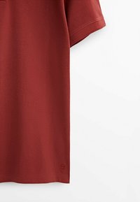 Polo à manches courtes rouge brique montrant un tissu texturé et un petit logo brodé près de l’ourlet du côté droit.