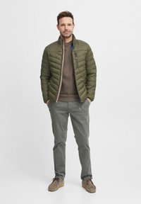 Olivengrüne Pufferjacke mit gestepptem Muster, Reißverschluss vorne und blauen Akzenten, kombiniert mit einem braunen Pullover und grauen Hosen. Beigefarbene Schuhe sind zu sehen.