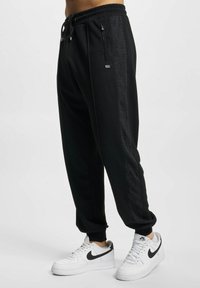  svarte joggebukser med snor i livet, smale ankler og sidelommer med glidelås. Har et mønstret stripe detaljer langs benet.