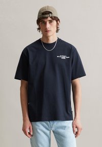 Marineblauwe katoenen t-shirt met korte mouwen. Voorzien van witte tekst op de borst. Gecombineerd met lichtblauwe denim jeans. Eenvoudig ontwerp.