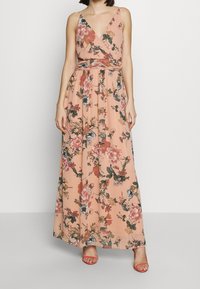 Maxiklänning i persikofärgad tyg med blommönster, V-ringning och justerbara axelremmar. Har en figurnära midja och en svängdkjol. Kombineras med röda sandaler.