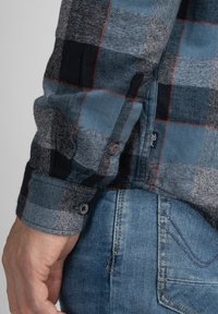 Camisa a cuadros en azul y gris con patrón de pata de gallo y acentos oscuros a cuadros en contraste. Presenta puños con botones y una etiqueta en el dobladillo.