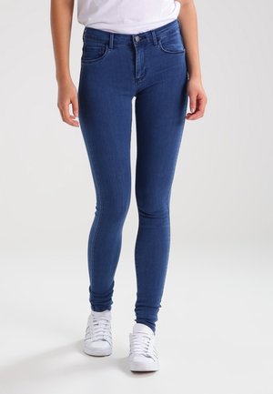 Person, die blaue Skinny-Jeans und weiße Sneakers trägt, steht vor einem schlichten hellen Hintergrund.