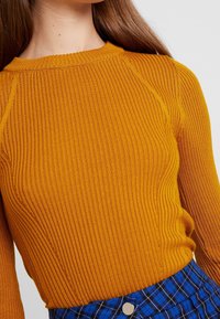 Senapsgul ribbad tröja med långa ärmar och rundad halsringning, med ett strukturerat mönster och en figursydd silhuett.